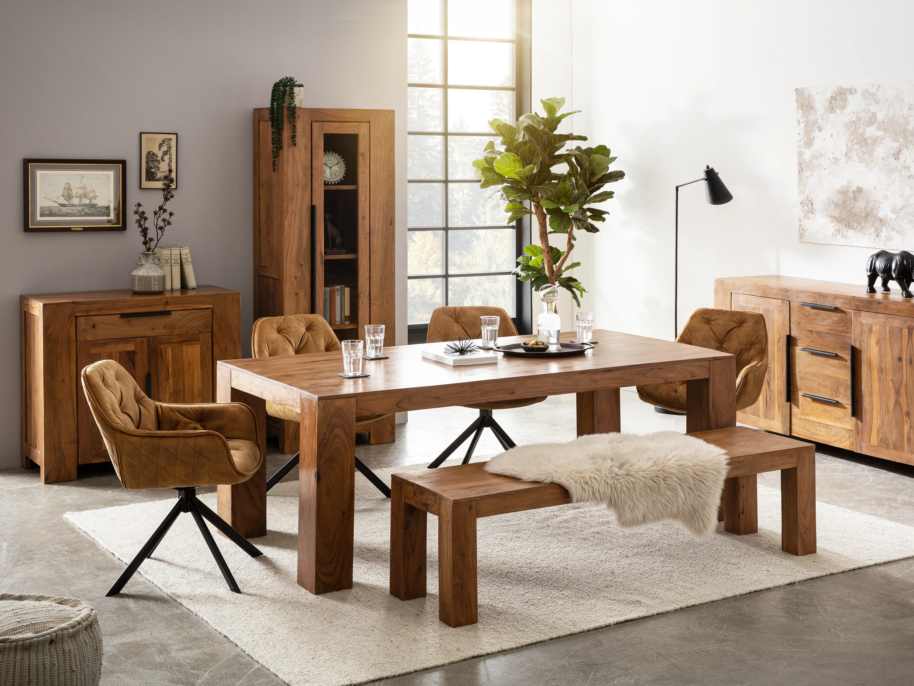 Essbereich mit modernem Holztisch, gepolsterten Stühlen und Bank auf hellem Teppich. Links steht die Kommode Osbon mit zwei Türen und einer Schublade aus braun lackierter Akazie. Oberflächen dekoriert mit Pflanzen und Bildern.